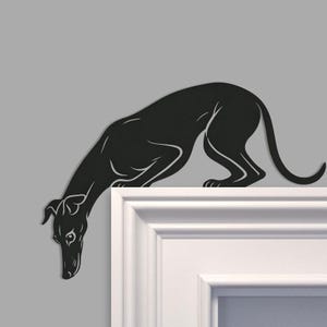 May include: A black metal silhouette of a Greyhound dog, with its head lowered. The dog is positioned on a white door frame against a grey background. The artwork captures the elegant form of the breed.