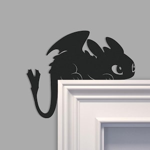 May include: A black dragon silhouette, featuring large wings and a curved tail, is positioned on a white surface against a gray backdrop. The dragon has large eyes and a friendly expression. This decorative piece is ideal for a child's room or a fan of fantasy creatures.