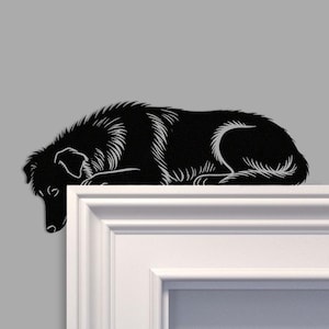 Puede incluir: Silueta de perro de metal negro, con detalles blancos, tumbada sobre una superficie blanca. El perro está representado en una postura de descanso, con la cabeza baja. El fondo es gris liso. El pelaje del perro está detallado.