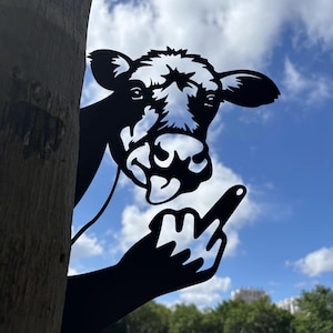 May include: Black metal cow silhouette with a hand giving the middle finger. The cow's head peeks out from behind a tree trunk, with a blue sky and clouds in the background. A humorous garden decoration.