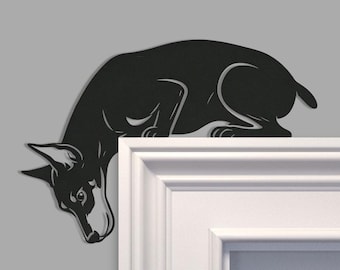 Puede incluir: Una pieza de arte mural de metal negro con forma de perro en posición agachada, diseñada para montarse en la esquina superior del marco de una puerta. La silueta presenta detalles recortados que definen las orejas, el hocico y la postura del perro.