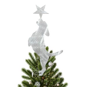 Op de afbeelding: Een zilveren hondvormige kerstboomtop die naar een zilveren ster reikt. De hond staat bovenop een groene kerstboom versierd met ornamenten en lichtjes. Een feestelijke vakantieversiering.
