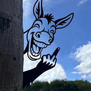 Könnte beinhalten: Schwarze Metall-Silhouette eines grinsenden Esels mit erhobenem Mittelfinger. Der Esel lugt hinter einem Baumstamm hervor, vor blauem Himmel mit Wolken. Humorvolle Gartendekoration.