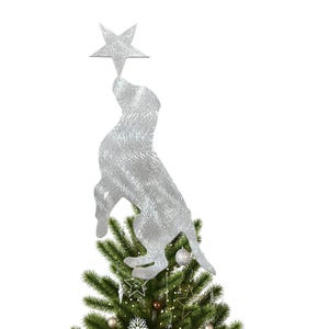 May include: A silver-colored dog-shaped Christmas tree topper reaching for a silver star. The dog is positioned above a decorated evergreen tree with ornaments and lights, creating a festive holiday scene.
