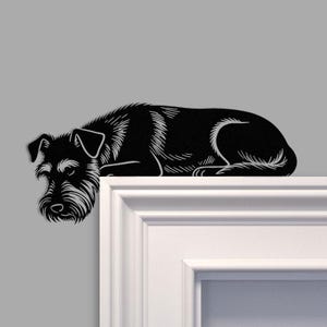 Pode incluir: Silhueta metálica a preto e branco de um cão Schnauzer, deitado. O cão está a descansar numa superfície branca, com a cabeça e as patas dianteiras penduradas na borda. O pelo do cão é detalhado com linhas brancas.