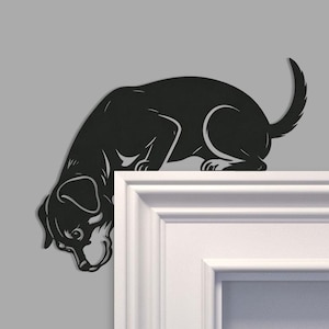 Könnte beinhalten: Schwarze Metall-Hunde-Silhouette, die über einen weißen Vorsprung zu blicken scheint. Der Hund befindet sich in einer nach unten gerichteten Pose, wobei sein Kopf und seine Vorderpfoten über den Rand hinausragen. Der Schwanz des Hundes ist nach oben gebogen. Der Hintergrund ist einfarbig grau.