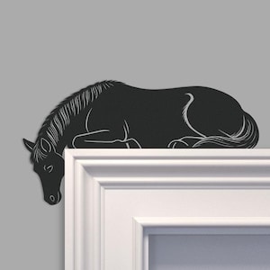 Puede incluir: Silueta de caballo de metal negro, descansando sobre una superficie blanca. El caballo se representa en una posición relajada, acostada, con su cabeza y crin detalladas en líneas blancas. La obra de arte está sobre un fondo gris.