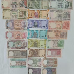 May include: A collection of Indian Rupee banknotes, featuring denominations from 1 to 1000. The notes display portraits of Mahatma Gandhi and various designs, with text in both English and Hindi. The banknotes are arranged on a white surface.