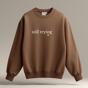 May include: A brown crewneck sweatshirt with the words "still trying" in white script and a small gold star. The sweatshirt has long sleeves and a ribbed collar, cuffs, and hem. The garment is made of a soft material.