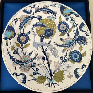 May include: A white ceramic decorative plate with a detailed floral and peacock design. The plate features a central peacock perched on a branch, surrounded by blue, pink, and green floral patterns. The plate is approximately 12 inches in diameter.