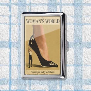 May include: A silver cigarette case with a graphic. The graphic shows a black high-heeled shoe and the text "It's a Woman's World" and "You're just lucky to be here."