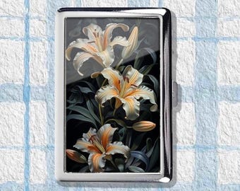 May include: A silver cigarette case featuring a floral design of white and orange lilies against a dark background. The lilies are in full bloom, with detailed petals and green leaves.