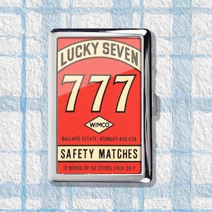 May include: A rectangular metal matchbox with a red background and the text "LUCKY SEVEN 777" in white and cream colors. The box also features the text "SAFETY MATCHES" and "WIMCO".