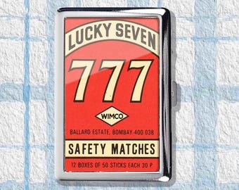 Puede incluir: Una caja de cerillas rectangular de metal con fondo rojo y el texto "LUCKY SEVEN 777" en blanco y crema. La caja también presenta el texto "SAFETY MATCHES" y "WIMCO".