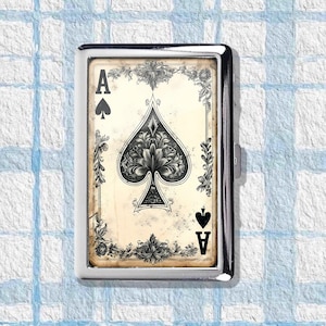 May include: A silver cigarette case featuring the Ace of Spades playing card design. The card has a black spade symbol and the letter "A" in the corners. The card is framed by floral and leaf patterns.