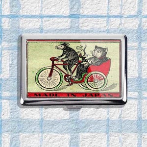 Puede incluir: Un estuche de cigarrillos de metal plateado con una colorida ilustración de una rata montando en bicicleta, tirando de un gato en un carruaje rojo. La caja tiene un borde rojo y las palabras "MADE IN JAPAN".