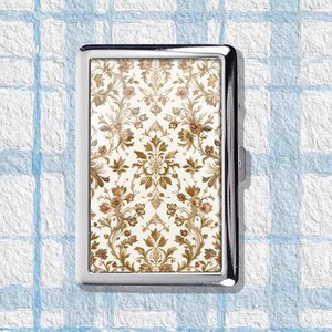 May include: A rectangular, silver-toned cigarette case. The case features a decorative, floral pattern in shades of brown, beige, and pink against a cream background. The design is symmetrical and ornate, with a classic, vintage aesthetic.