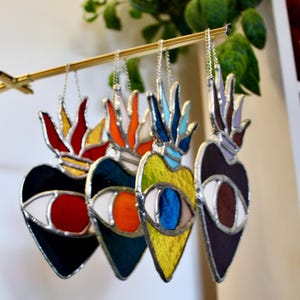 Peut inclure: Quatre ornements en forme de cœur en vitrail avec des motifs d'œil sont suspendus à une barre dorée. Chaque cœur présente une palette de couleurs différente : noir avec rouge, sarcelle avec orange, jaune avec bleu et violet avec marron. Les ornements ont des chaînes argentées.