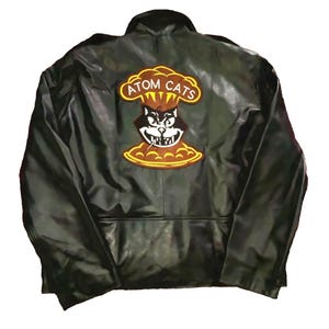 Può includere: Giacca in pelle nera con il logo Atom Cats sul retro. Il logo presenta la faccia di un gatto all'interno di un design a fungo atomico con le parole "ATOM CATS" in giallo.