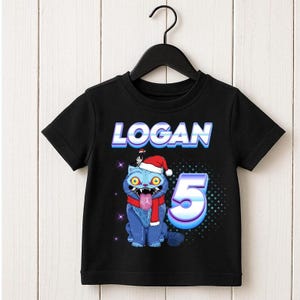 May include: Black t-shirt featuring the name "LOGAN" in a blue and white font. A blue cartoon cat wearing a Santa hat and scarf is below the name, with the number "5" next to it. The cat has its tongue out.