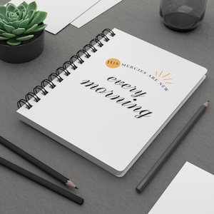 Op de afbeelding: Een wit notitieboek met spiraalbinding en de tekst His mercies are new every morning in zwarte en oranje letters. De kaft bevat een kleine oranje cirkel en een zonnestraalgrafiek. Twee zwarte potloden liggen in de buurt op een donkergrijs oppervlak.