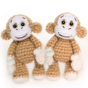 Może przedstawiać: Dwie ręcznie robione szydełkowe małpy. Każda małpa ma brązowy tułów, białą twarz, dłonie i stopy oraz brązowe uszy. Mają czarne oczy i wyszyty uśmiech. Małpy stoją wyprostowane, obok siebie.