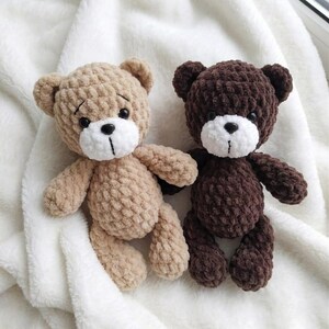 May include: Two handmade crochet teddy bears. One is light brown, the other is dark brown. Both have white muzzles, black eyes, and stitched mouths. They are lying on a soft, white, textured blanket.