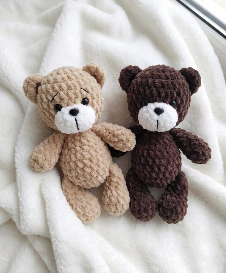 May include: Two handmade crochet teddy bears. One is light brown, the other is dark brown. Both have white muzzles, black eyes, and stitched mouths. They are lying on a soft, white, textured blanket.
