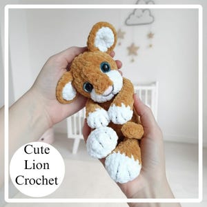 May include: A handmade crochet lion plush toy in shades of brown and white. The lion has large blue eyes, a pink nose, and white accents on its ears and paws. The text "Cute Lion Crochet" is in a white circle.