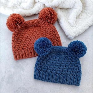 Puede incluir: Dos gorros de ganchillo, uno de color naranja quemado y otro de color azul intenso, cada uno con dos grandes pompones a juego en las esquinas superiores. Ambos gorros tienen un patr&oacute;n de punto texturizado y un borde acanalado, con un dise&ntilde;o uniforme.