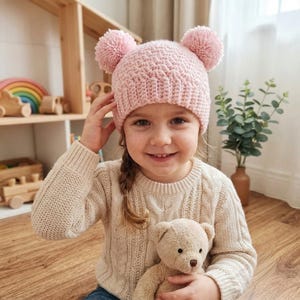 Puede incluir: Un gorro de ganchillo de color rosa claro que presenta dos grandes pompones a juego en la parte superior. El gorro est&aacute; confeccionado con un patr&oacute;n de punto texturizado y un borde acanalado que se ajusta a la cabeza con suavidad.