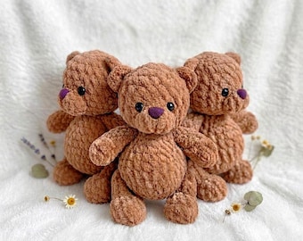 Puede incluir: Tres osos de peluche tejidos a ganchillo con hilo marrón suave y afelpado. Cada oso presenta pequeños ojos de plástico negro y una distintiva nariz bordada en color morado. Los osos están dispuestos juntos, mostrando su textura.