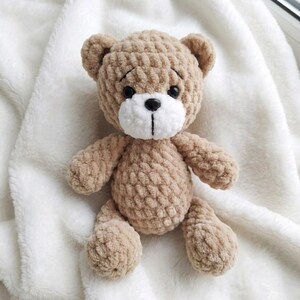 May include: A handmade, light brown teddy bear with a white muzzle and black eyes. The plush toy is sitting on a white, fluffy blanket. The bear has a classic design with a round head, small ears, and a stitched nose.