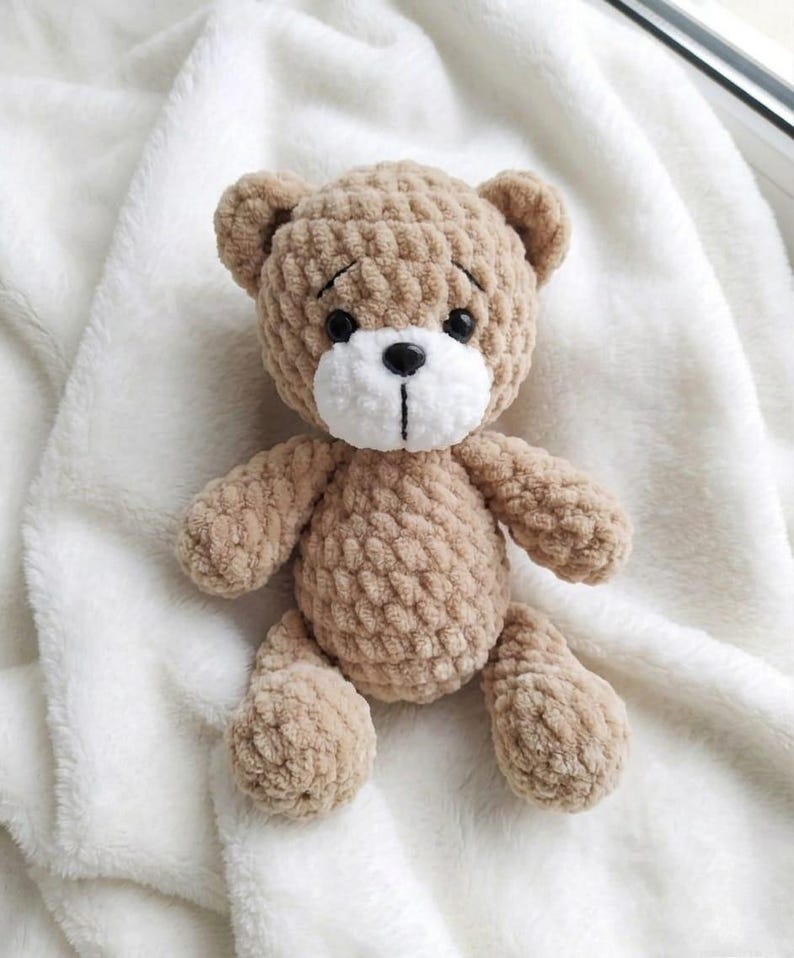 May include: A handmade, light brown teddy bear with a white muzzle and black eyes. The plush toy is sitting on a white, fluffy blanket. The bear has a classic design with a round head, small ears, and a stitched nose.