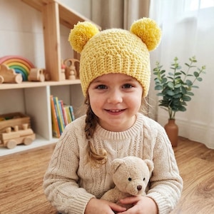 Puede incluir: Un gorro de ganchillo de color amarillo brillante con dos grandes pompones en la parte superior. El gorro tiene un borde acanalado texturizado y un patr&oacute;n de puntos denso y uniforme en toda la corona, ofreciendo un aspecto suave y acogedor.