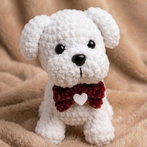 Pode incluir: Um cachorrinho branco feito à mão em crochê, com olhos e nariz pretos, e uma gravata borboleta bordô com um detalhe em forma de coração branco. O brinquedo de pelúcia está sentado em uma superfície marrom macia.