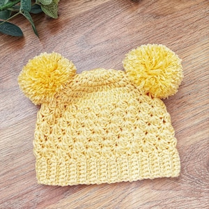 Puede incluir: Un gorro de ganchillo amarillo brillante que presenta un patr&oacute;n de punto de concha texturizado en todo el cuerpo, un borde acanalado en la base y dos pompones de hilo grandes y esponjosos unidos a las esquinas superiores de la corona.