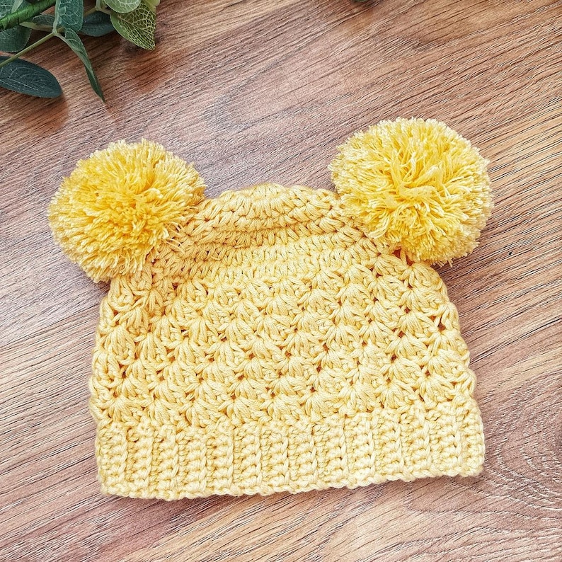Puede incluir: Un gorro de ganchillo amarillo brillante que presenta un patr&oacute;n de punto de concha texturizado en todo el cuerpo, un borde acanalado en la base y dos pompones de hilo grandes y esponjosos unidos a las esquinas superiores de la corona.