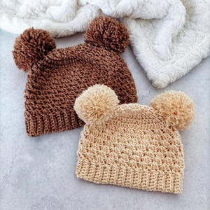 Puede incluir: Dos gorros tejidos a ganchillo con pompones en la parte superior. Un gorro es de color marr&oacute;n oscuro y el otro es de color beige claro. Ambos gorros presentan un patr&oacute;n de puntos texturizado y repetitivo con un borde acanalado a juego.