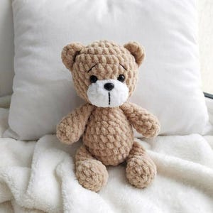 May include: A handmade, tan-colored teddy bear with a white muzzle and black eyes. The bear is crocheted with a textured yarn and has a stitched black mouth. It is sitting on a white, fluffy blanket.