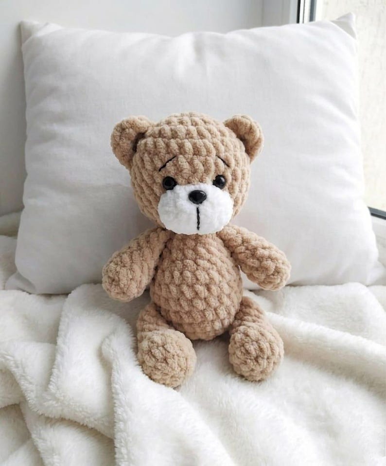 May include: A handmade, tan-colored teddy bear with a white muzzle and black eyes. The bear is crocheted with a textured yarn and has a stitched black mouth. It is sitting on a white, fluffy blanket.
