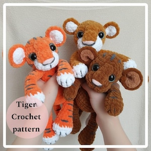 May include: Three crochet tiger cubs in shades of orange and brown are held up. The orange cub has black stripes, white paws, and large black eyes. The brown cubs have white accents. The text "Tiger Crochet pattern" is in a pink circle.