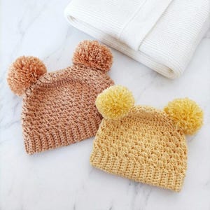 Puede incluir: Dos gorros de ganchillo con pompones, uno en tono marr&oacute;n claro y otro en tono amarillo mostaza. Ambos gorros presentan un patr&oacute;n de puntos texturizados y un borde acanalado. Una tela blanca de punto gofrado doblada se sit&uacute;a en la esquina.