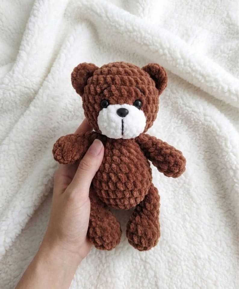 May include: A handmade brown teddy bear with a white muzzle and black eyes. The bear is made of a textured, plush material and has a classic teddy bear shape. The bear is a soft toy.