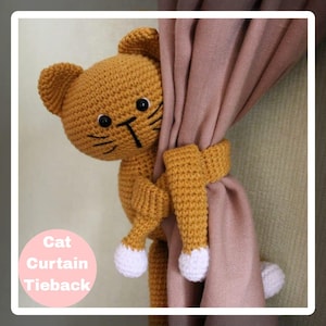 Puede incluir: Un recogedor de cortinas de gato tejido a ganchillo en tonos naranja y blanco. El gato tiene ojos y bigotes negros y sujeta una cortina. El texto "Cat Curtain Tieback" está en un círculo rosa.