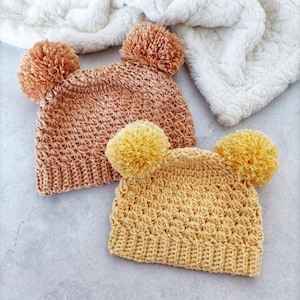 Puede incluir: Dos gorros de ganchillo con un patr&oacute;n de punto de estrella texturizado y bordes acanalados. Un gorro es de color beige c&aacute;lido con dos pompones a juego, y el otro es de color amarillo brillante con dos pompones amarillos a juego arriba.