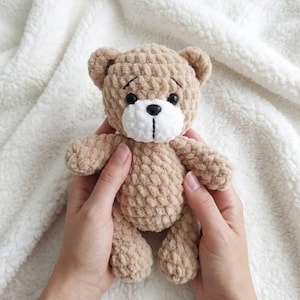 May include: A handmade, tan crochet teddy bear with a white muzzle and black eyes. The bear is held in two hands against a white, textured background. The bear's features include small ears, a stitched nose, and a slightly sad expression.
