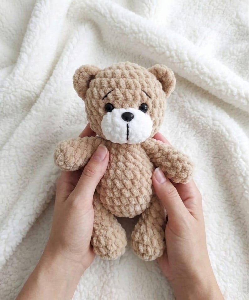 May include: A handmade, tan crochet teddy bear with a white muzzle and black eyes. The bear is held in two hands against a white, textured background. The bear's features include small ears, a stitched nose, and a slightly sad expression.
