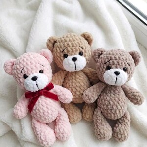 May include: Three handmade crochet teddy bears in shades of pink, tan, and brown. Each bear has a white muzzle, black eyes, and a stitched mouth. The pink bear has a red velvet bow. The bears are displayed on a white, textured surface.