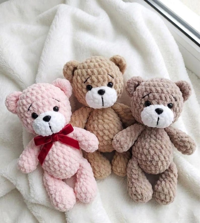 May include: Three handmade crochet teddy bears in shades of pink, tan, and brown. Each bear has a white muzzle, black eyes, and a stitched mouth. The pink bear has a red velvet bow. The bears are displayed on a white, textured surface.